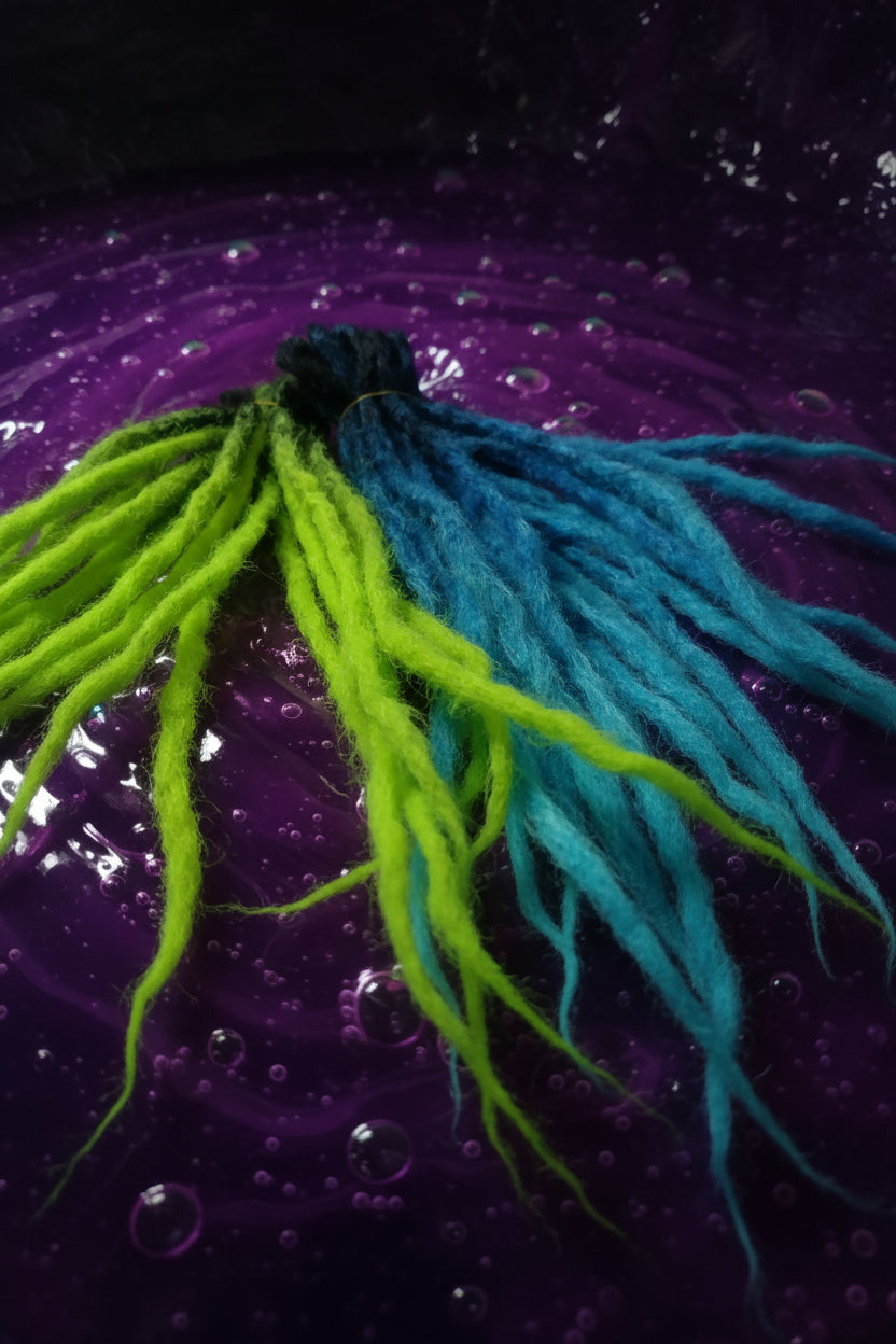A close-up of a hairpiece or dreadlock extension with bright neon green and teal-blue synthetic fibers gathered at the top, shown against a pink fabric background.