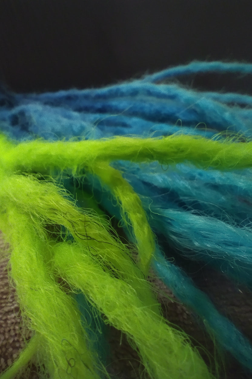 Close-up of fluffy yarn strands in bright lime green and medium blue, held in a hand. A vertical watermark reads 'October Serenity Products' on the right side.